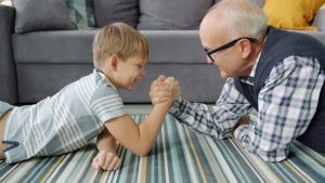 Elderly man and little boy are practising arm wresting at home enjoying activity, child is winning. Modern pastime, family relationship and fun concept.